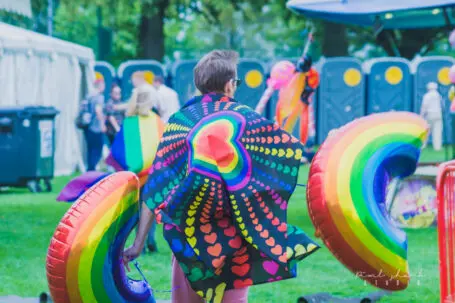 Person from behind wearing a colourful rainbow heart cape holding a rainbow shaped balloon in each hand