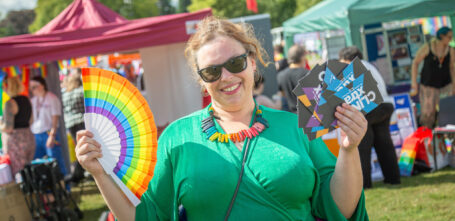 Vibrant, smiling person holding leaflets and a rainbow pride fan at a Pride event